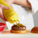 A hand holding a Vollrath Tri Tip Squeeze Bottle with yellow cap pouring mustard onto a burger.