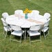 A Lancaster Table & Seating white plastic round folding table with white folding chairs and a vase of sunflowers.