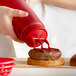 A hand using a Vollrath red Tri Tip squeeze bottle to pour ketchup onto a burger.