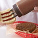 A person using a Vollrath brown Tri Tip wide mouth squeeze bottle to put ketchup on a hamburger.