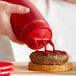 A person using a Vollrath Tri Tip squeeze bottle with red cap to pour ketchup on a burger.