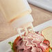 A clear squeeze bottle with a tri-tip nozzle dispensing sauce onto a sandwich.