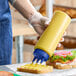 A clear 32 oz. squeeze bottle with a blue tri-tip cap being used to dispense a yellow condiment onto a slice of bread.
