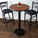 A Lancaster Table & Seating bar height table with a 24" round walnut table top and a cast iron base plate with a glass of beer on it.