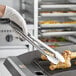 A person in gloves using Tablecraft locking tongs to pick up a chicken leg from a grill.