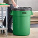 A man standing next to a Lavex green commercial trash can with a black bag in it.