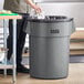 A man standing next to a Lavex gray round commercial trash can with a black plastic bag in it.