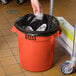 A person holding a Lavex orange plastic trash can with a silver lid.