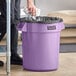 A person putting a black plastic bag in a purple Lavex 20 gallon round commercial trash can.