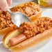 A person using a spoon to put Vanee Chili Hot Dog Sauce on a hot dog.
