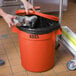 A hand putting a plastic bag into an orange Lavex trash can.