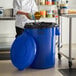 A woman in a chef's uniform putting food into a blue Lavex trash can.