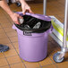 A person holding a purple Lavex commercial trash can lid over a trash can filled with foil.