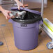 A hand putting foil in a Lavex purple round trash can with a lid.