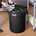A person's hands holding a plastic bag and putting it into a Lavex 32 gallon round black trash can.