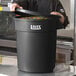 A man in a black chef's coat standing in front of a large Lavex black trash can with a lid open.