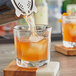 A person pouring orange liquid from a juicer into an Anchor Hocking Clarisse rocks glass with ice on a table.