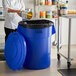 A woman pouring a can into a Lavex blue trash can in a school kitchen.