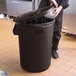 A person putting a plastic bag into a brown Lavex round commercial trash can.