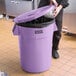 A person in black pants putting a plastic bag into a purple Lavex trash can with a lid.