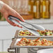A person holding a pair of American Metalcraft stainless steel tongs over food on a buffet table.