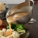 A Tablecraft stainless steel gravy boat pouring gravy over a plate of meat and potatoes.