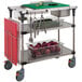 A Metro PrepMate MultiStation kitchen cart with stainless steel shelving holding a plastic container of onions and utensils.