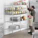 A man in a brown apron standing in front of a Regency metal wire shelf with cans on it.