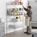 A man in a grey shirt and apron standing in a professional kitchen with a Regency chrome wire shelf full of canned food.