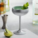 A silver coupe glass filled with a drink and lime wedges.