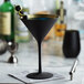 A close-up of a black Stolzle Glisten martini glass with a gold rim filled with a blue liquid and olives.