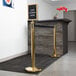 A Lancaster Table & Seating gold crowd control stanchion with a sign that says "Line Forms Here" and clear covers.