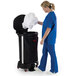 A woman in blue scrubs using a Rubbermaid black linen hamper to put a white shirt inside.