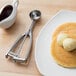 A metal ice cream scooper with a squeeze handle on a table with a plate of pancakes and ice cream.