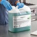 A person wearing blue gloves holding a large plastic container of green liquid.