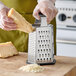A six-sided stainless steel box grater with a soft grip handle being used to grate cheese.