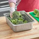 A person holding a piece of broccoli in a Choice 4" deep stainless steel pan.