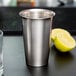 A close-up of a Vollrath stainless steel cup with a lemon on a black surface.