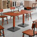 A rectangular solid wood live edge table top with a rustic mahogany finish, supported by two metal bases, surrounded by matching wooden chairs in a dining area.
