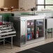 An Avantco stainless steel back bar refrigerator on a metal shelf filled with bottles.