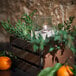 A Sterno Pinot votive candle in a glass holder with green leaves and an orange on a table.