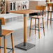 A Lancaster Table & Seating bar height table with two wooden stools and wine glasses on it.