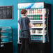 A woman in a blue shirt standing in front of a black Avantco refrigerated air curtain merchandiser.