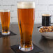 Two Libbey plastic Pilsner glasses of beer on a table with a bowl of nuts.