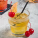 A Libbey Tritan plastic rocks glass with yellow liquid, ice, and orange and cherry garnish.