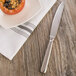 An Oneida Baguette stainless steel dessert knife on a white napkin next to a plate of food.