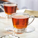 Two Acopa glass cups of tea on saucers with sugar cubes on a table.