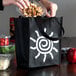 A person putting a plastic container into a black non-woven shopping bag with a sun design.