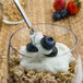 A Fineline Tiny Tasters silver plastic cocktail spoon in a glass of yogurt with blueberries.