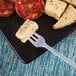 A Fineline Tiny Tasters silver plastic cocktail fork spearing a piece of cheese on a plate.